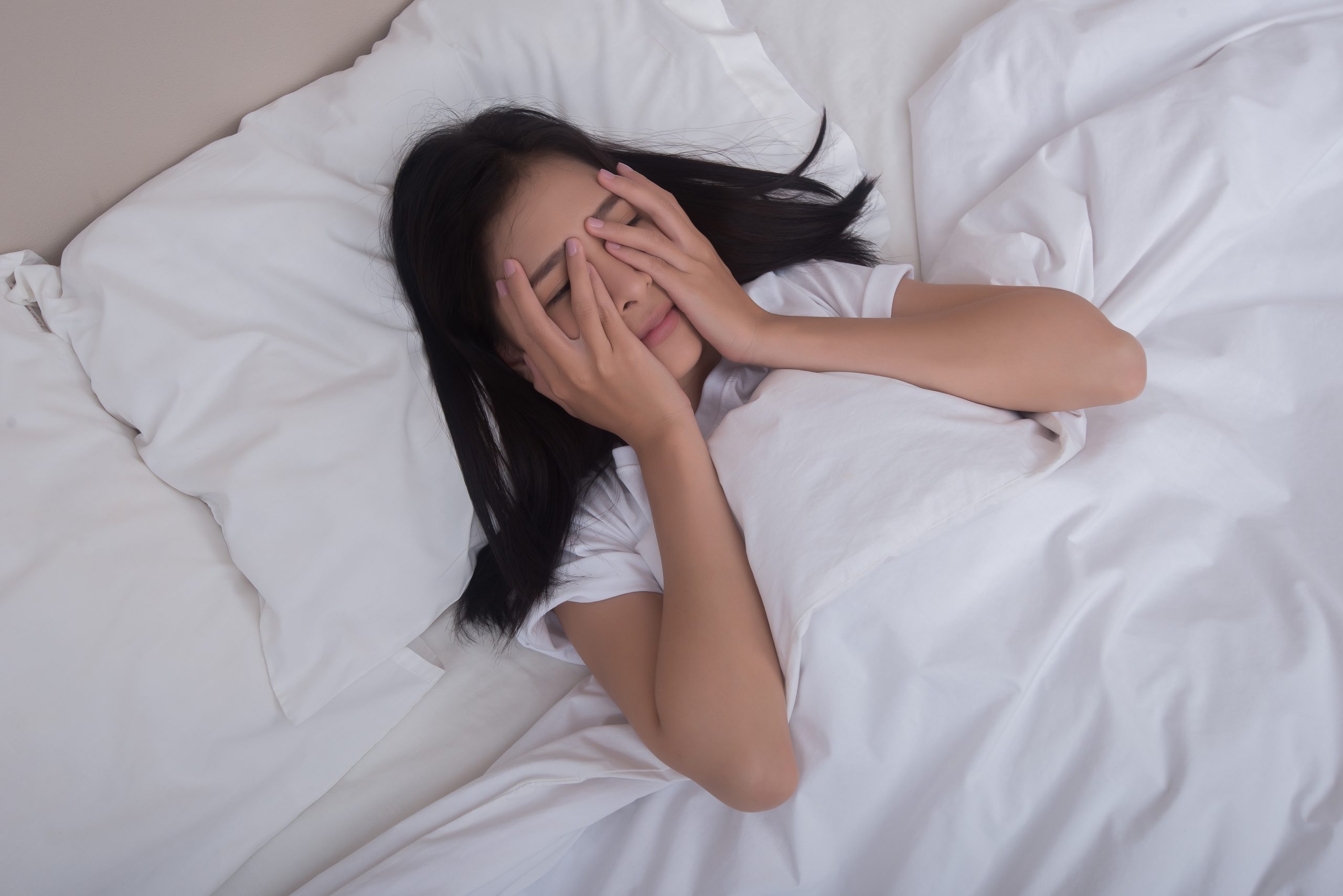Beautiful young woman covering her eyes, looking at camera and smiling while lying in the bed