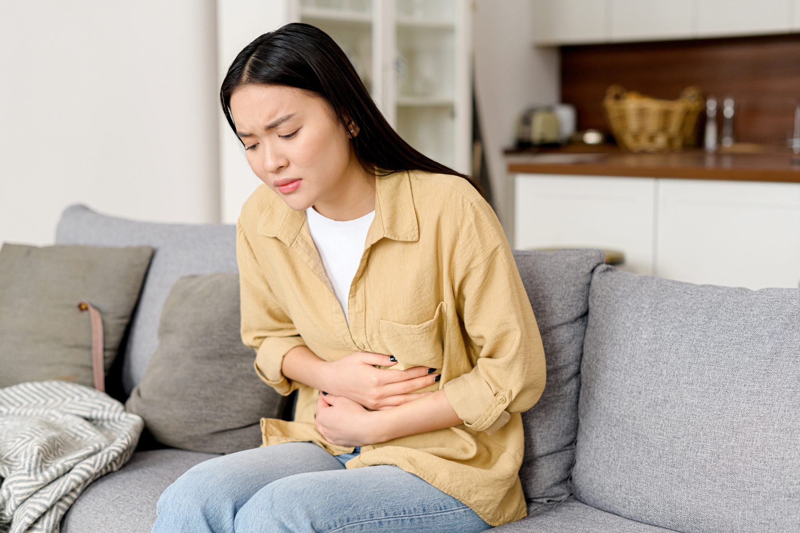 Concerned asian woman suffering from stomach-ache sitting on sofa. Female holding tummy, undergoing belly pain and discomfort, suffer from menstruation